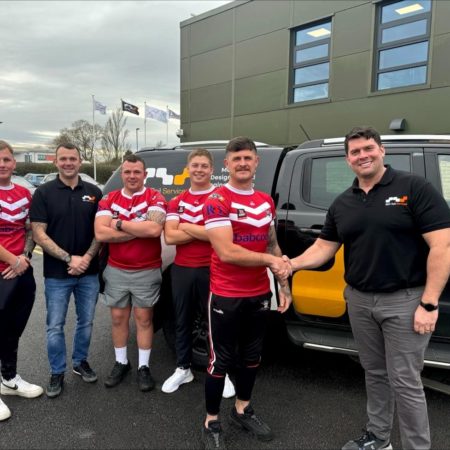 Group of men posing in sports jerseys alongside a branded vehicle, with two in black shirts shaking hands.